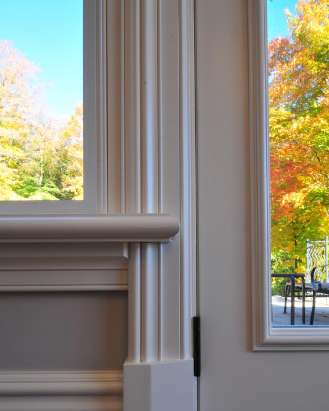 White decorative wood trim and moulding combination framing a window and doorway with autumn trees visible outside.